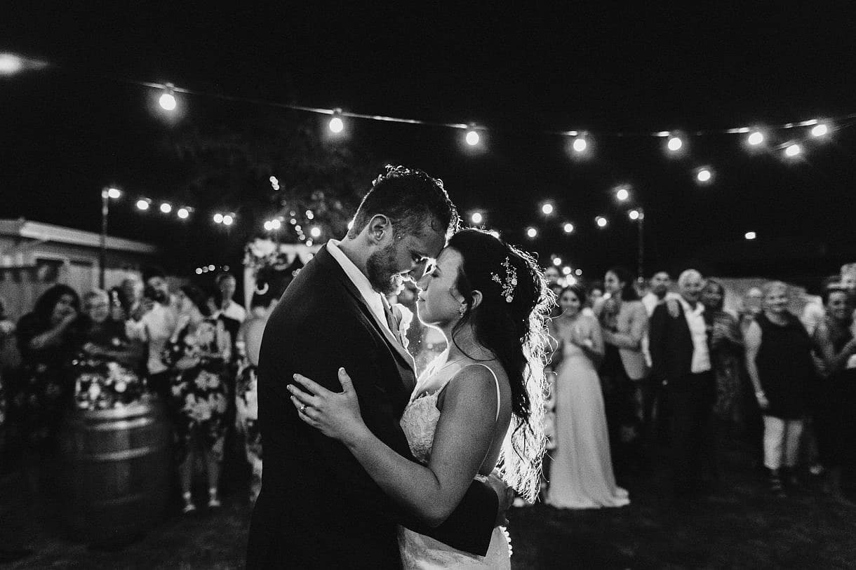 Wedding couple dance under lights and with family & friends glowingly supporting them in the background